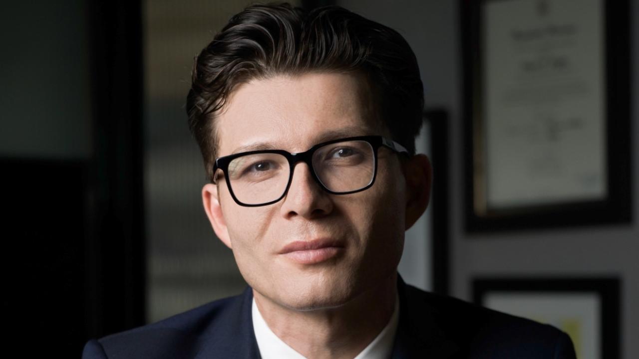 A man in a suit and glasses sits confidently at a desk, hands clasped, against a professional backdrop.