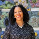 A smiling woman with dark curly hair, wearing a dark polka-dot shirt and a chunky white necklace, standing in front of a colorful mosaic wall featuring flowers and insects